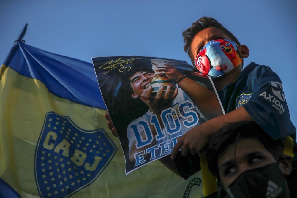 A kid sitting on the shoulders of a man holds a poster of Maradona in front of the Casa Rosada presidential palace, as people gather to mourn the death of soccer legend Diego Armando Maradona, in Buenos Aires, Argentina, November 26, 2020. REUTERS/Ricardo Moraes
