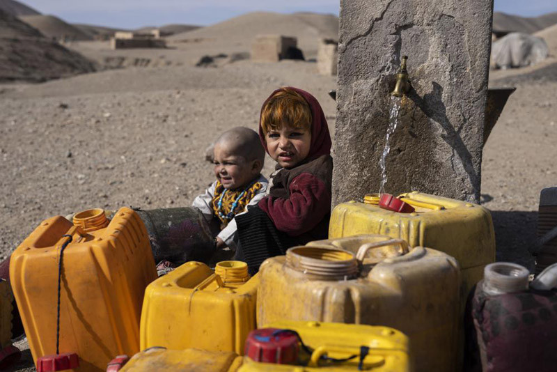 Two Afghan children sit next to a spigot as people of Kamar Kalagh village outside Herat, Afghanistan, try to fill their plastic containers with water, on Friday, Nov. 26, 2021. Afghanistan’s drought, its worst in decades, is now entering its second year, exacerbated by climate change. (AP File Photo)