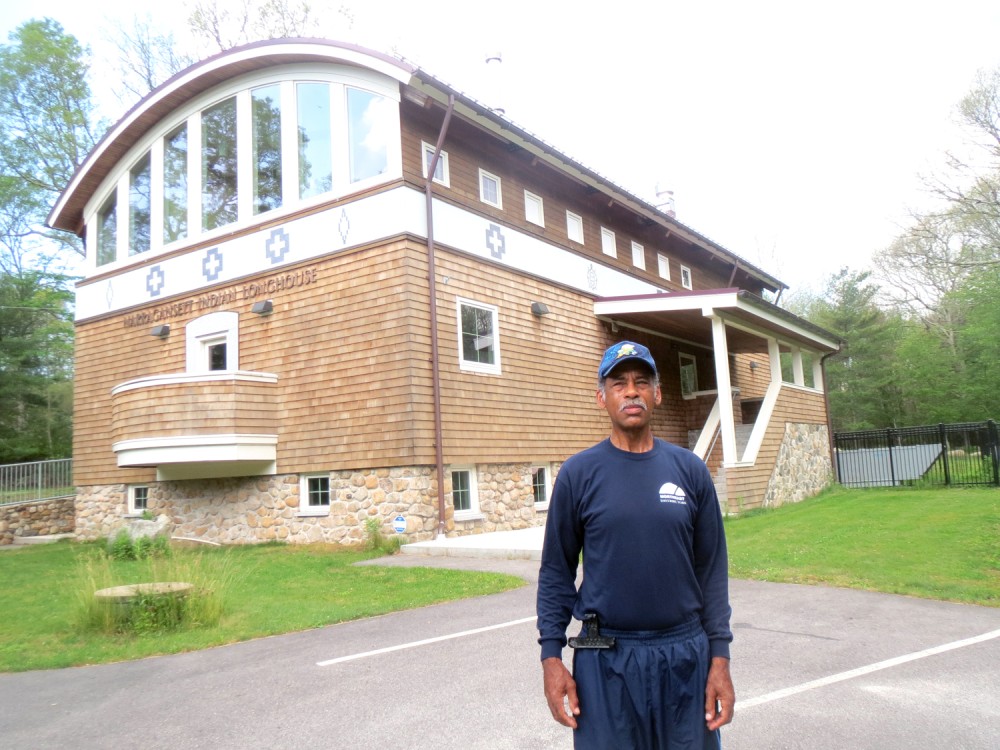 Narragansett elder Hiawatha Brown (63) stands tall in front of the Narragansett Indian Longhouse on their reservation in Rhode Island (USA) where important meetings of his people are held. The Longhouse (a sort of Morung) was designed by his mother, the medicine woman. Its construction was completed, with modern finishing, five years back. The crosses on the body of the structure signify the four earthly directions—east, west, north and south. The other symbols denote clans.  (Morung Photo)